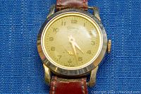 Close-up of the GENOVA watch face showing the gold-tone round case, beige dial with gold Arabic numerals and hands. Scratches visible on glass.