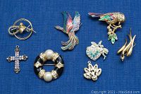 Seven costume jewelry pieces displayed on a blue cloth background, showing brooches and a pendant with various colors and metal finishes.