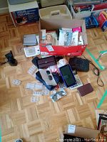 Photo showing Omron blood pressure monitor with cuff, new ladies underwear, wallets, pill boxes, GE clock radio, calculators, and other small items scattered on wood floor.