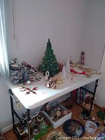 Photo showing large green ceramic tree with colorful bulbs and additional assorted Christmas ornaments on a white table near a window.