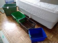 View of all four bins arranged in a line on the floor showing various sizes and colors (green, black, blue) next to a bed.