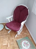 Front view of white wooden glider chair with burgundy padded cushions, placed on parquet floor near window with blinds.