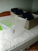 Four large beige plastic storage bins each topped with a black lid, placed on a bed.