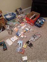 Wider view of assorted tools, hardware, and painting supplies laid out on carpet including Black and Decker drill body without battery, hand saw, hammer, stapler, drill bit sets, screws, peg hooks, switch plates.