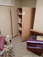 Photo of a light brown faux wood cabinet with two open doors showing shelves inside, placed in a basement corner near a floral chair and stacked linens.