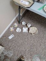 Overview of lot items on carpet floor next to table leg including porcelain dishware and decorative items.