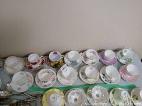 Wide view of 15 assorted floral teacups and matching saucers laid out on table showing various patterns and colors.