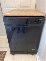 Front view of black Whirlpool portable dishwasher with maple wood top on wheels.