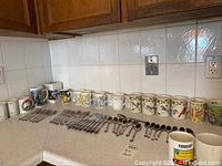 Over 24 assorted mugs arranged in a row with 37 pieces of matching flatware (forks, spoons, and knives) laid out in front.