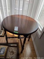 Top view of round wooden drop leaf table with label showing 27X27X27 dimensions in inches, some wear visible on surface.