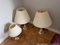Three table lamps arranged on parquet flooring against a wall, showing two taller silver-based lamps and one smaller gold-based lamp with cream pleated shades.