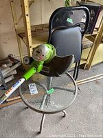 Photo showing the GreenWorks electric string trimmer, patio glass side table, and black folding chair, all arranged together in a storage setting.