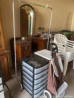 Metal coat rack, plastic storage drawer unit, wheeled shopping tote, and stacked white plastic folding chairs in garage setting.