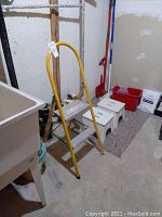 Yellow metal step ladder with two steps and handrails, shown in basement environment with concrete floor and walls.