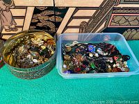 Two containers filled with assorted vintage jewelry pieces including brooches, necklaces, earrings, and pendants in various styles and colors.