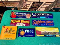 Seven colorful vintage crate labels for tomatoes, grapes, and other produce laid out on green fabric.