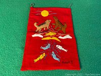 Full view of vintage red fabric Inuit tapestry showing two wolves, fish, the sun, and snow patches with embroidered details and signature.