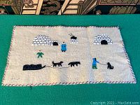 Full view of the white wool tapestry with needlework of igloos, sled dogs, sled, and figures dressed in parkas.