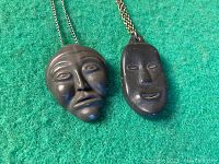 Two vintage Inuit soapstone face pendants lying side by side on green fabric background showing detailed face carvings and metal chains.