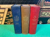 Three hardcover books standing upright, blue, green, and red cloth covers with gold embossed lettering and decorative designs on spines.
