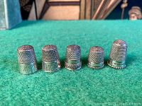 Front view of five silver thimbles on green felt, showing varying designs and sizes.
