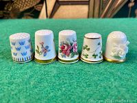 Frontal and angled view of five vintage fine bone china thimbles in detailed decorative styles on a green felt surface.