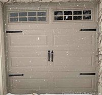 Front view of installed beige sectional garage door with black decorative hinges, handles, and a row of eight windows across the top panel. Snow on driveway.