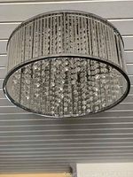 Photo showing a round flush mount ceiling light fixture mounted on a white paneled ceiling. The fixture has a metal frame with vertical rods and interior hanging glass or crystal beads.