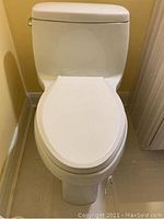 Front view of white ceramic two-piece toilet with elongated bowl and seat installed against yellow wall