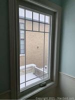 Vinyl casement window installed in wall with visible white frame, decorative grid on glass, crank handle at bottom and trim surrounding frame