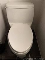 Front-facing image of a white ceramic toilet with an oval seat and a matching tank in a tiled bathroom corner.