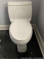 Photo of a white two-piece toilet, showing front view with closed lid and tank against wall.