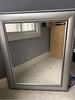 Photo showing a rectangular mirror with a broad silver frame with beaded accents, standing upright on the floor in front of a wall and window.