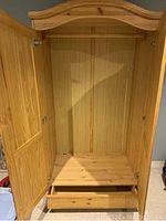 Interior view of the wooden armoire showing the hanging rod, shelf, and bottom drawer pulled out.