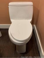 Front view of a white ceramic toilet installed on a floor with wood-like tiles against a peach colored wall.