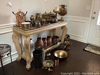 Wide view of all metal decor items on and under a marble-topped table in a foyer area, featuring copper ice bucket, kettles, pitchers, bowls, and plant stands.