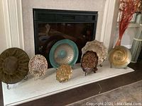 Seven decorative plates arranged on a mantle, showing variety in size, color and design with some plates on metal stands.