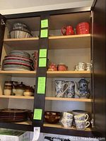Kitchen cabinet shelves displaying assorted plates, bowls, mugs, teapot with various patterns and colors including blue and white floral and brown glaze.
