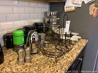 View of black ceramic canisters, metal salt and pepper shakers, metal two-tier basket, metal paper towel holder, and other kitchen items on granite countertop near white tile backsplash.