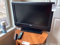26-inch Sanyo television placed on a wooden table near a window with remote and a battery by its side.