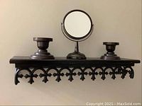 Photo of dark brown decorative wall shelf topped with a round standing mirror in the center and two broad-base candle holders on each side, all made of metal.