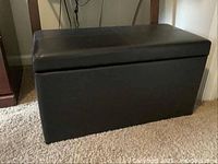 Front view of the brown vinyl storage bench with visible hinged lid, showing the bench on carpeted floor near a wooden table leg and wall outlet.