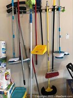 Photo showing several cleaning tools hanging on wall rack, including brooms, mops, yellow scrub bucket/dustpan, Hurricane spin broom. Surrounding area with paper towels and cleaning supplies visible.
