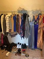 Photo of women's clothing hanging on a rack including white puffer coat, coats, scarves, evening dresses, and shoes and boots beneath the rack.