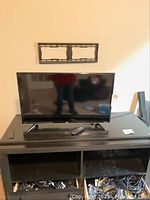 Front view of TCL Roku TV on black TV stand with remote on stand and wall mount bracket behind.