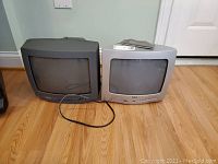 Two CRT televisions side by side on wood floor, one RCA in silver, the other Sanyo in grey