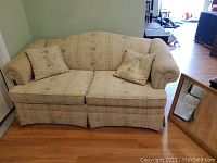 Front view of beige floral upholstered sofa with two matching throw pillows on wooden floor, against pale green walls.