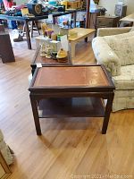 Two matching wooden side tables with reddish-brown leather inset tops edged with gold tooling, a lower shelf, and dark wood finish, set on a wooden floor.