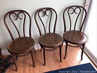 Photo showing three brown bentwood wooden chairs with curved backrest design, placed against a wall near a window on wooden floor. Shows overall condition and style.