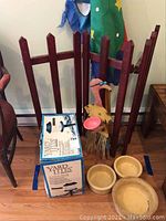 Wooden folding gate with metal flamingo decoration holding a pink dish, three tan ceramic dog bowls on floor, and boxed wired yard lights in front of the gate.